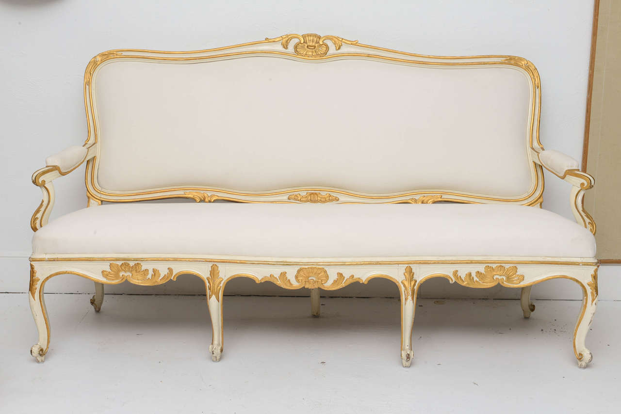 Swedish white settee with gilded ornaments and centered shell on the painted frame. Seat, back and elbow rests are upholstered in muslin.