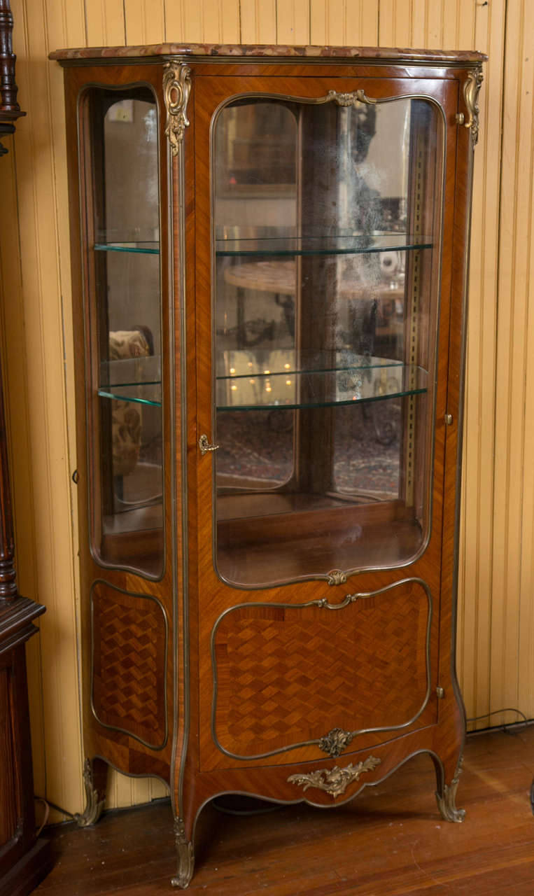 A very fine quality one door bronze mounted mahogany and parquetry vitrine, or curio cabinet, with serpentine front and sides.
It has two glass shelves, a beautiful marble top, and has had lights installed on top for viewing.The piece has the 