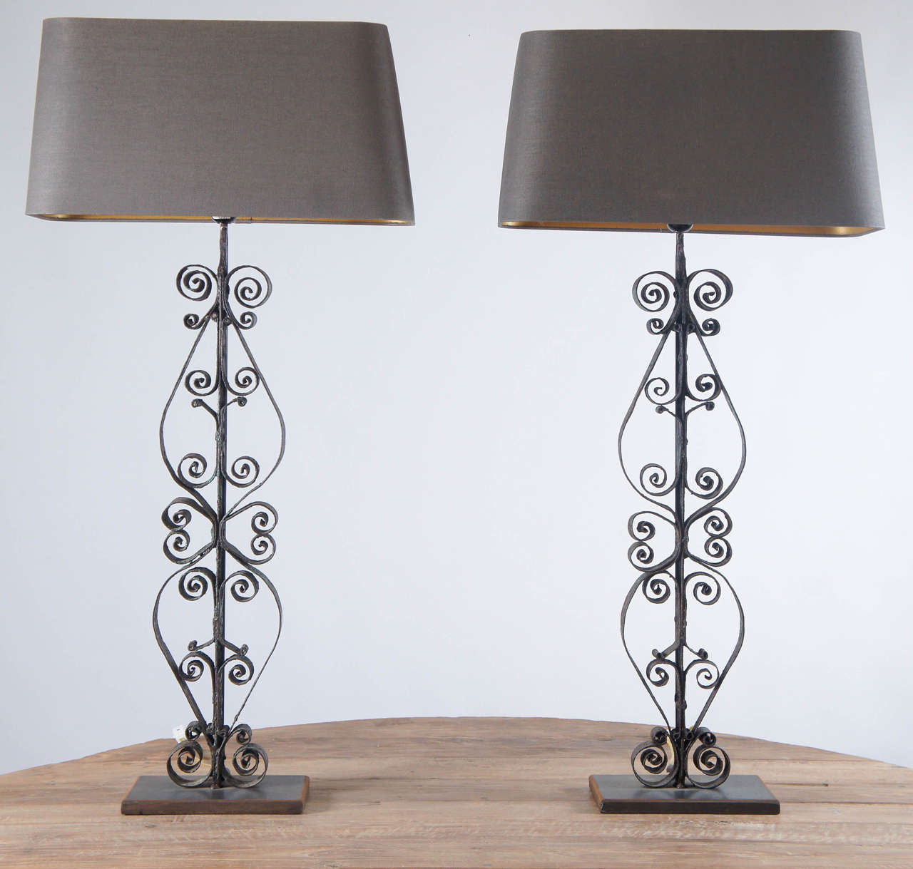 Pair of lamps composed of antique handmade wrought iron balusters and metal bases wired for US electrical use. The balusters are partially crusted with blue-green paint.  The rounded rectangular taupe shades are lined in gold to give off a warm