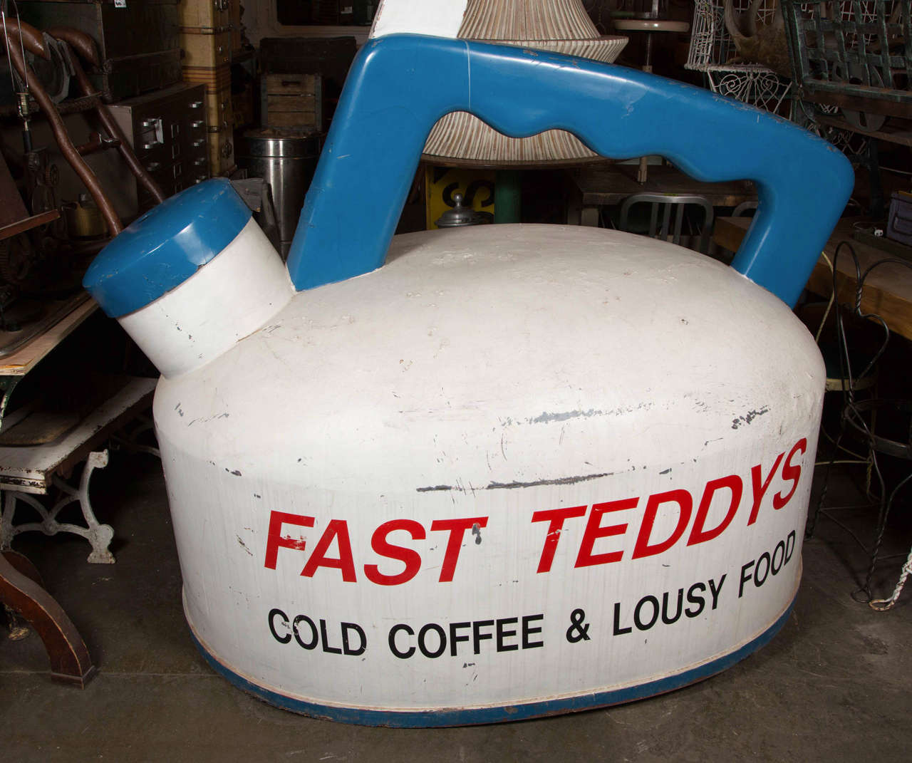 This is a larger than life 1940s Fast Teddy's Restaurant teapot sign. This sits flush to the ground with a blue handle and spout. The teapot itself is a scuffed white with red and black lettering. This can be seen at our 400 Gilligan St location in