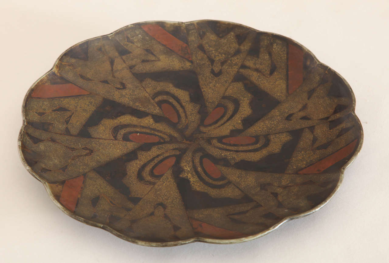 Of lobed form with wavy rim decorated with a geometric design of red, black and oxidized brass.

Signed: ''F. Grange''.