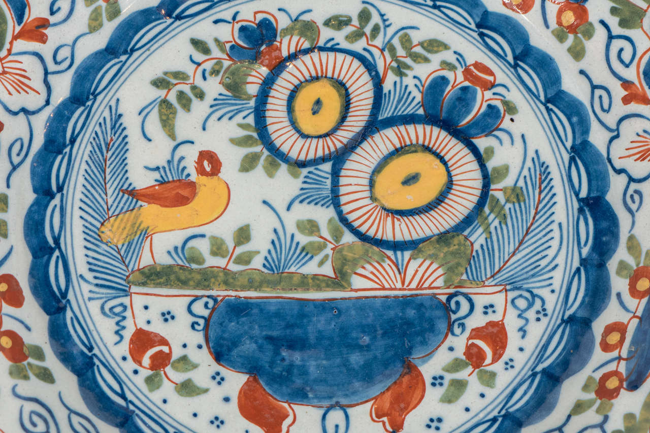 A Dutch Delft charger boldly painted in polychrome colors.The center shows a bird in a garden perched by a flower filled vase painted in bright orange, blues greens and yellows. Surrounded by a wide border of tulips and geometric shapes. .