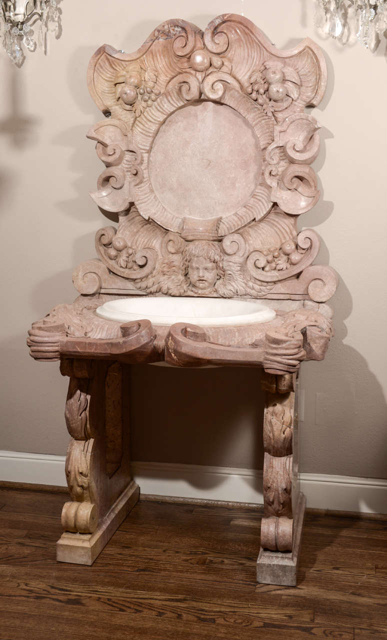 An exuberant carved lavabo with fruit, cherub's face and mythical masks.  Scrolls encircle a round cartouche where the water spout would go on the back panel. This consists of four separate pieces: two bracket leg panels, a carved deck with white