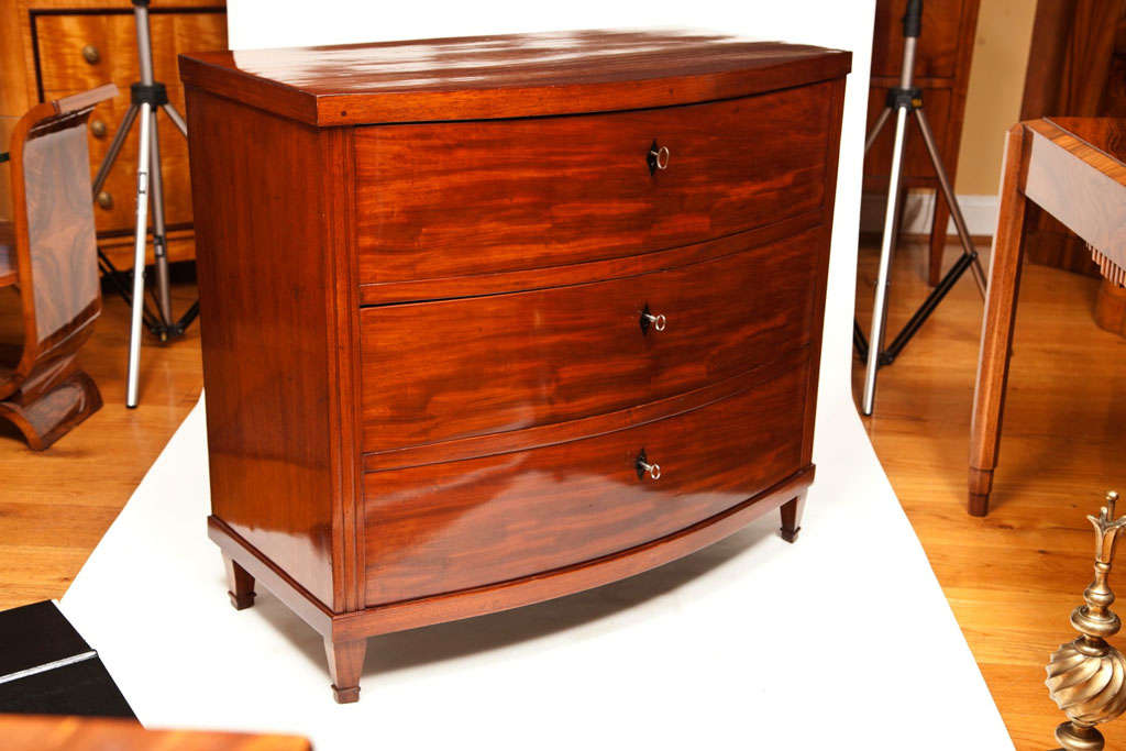 A pair of early Biedermeier mahogany bow-fronted chests comprised of three drawers with fluted front sides terminating on straight, tapered feet, adorned with ebony inlaid shield escutcheons