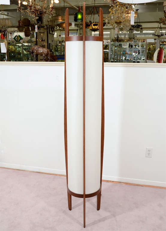A tall three-legged floor lamp in teak with a cylindrical, white shade.