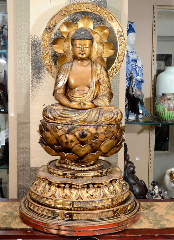 A large gilt wood sculpture of Buddha in traditional dhyana mudra pose of meditation. The figure contains much of the iconography inherent in depictions of the Buddha such as the lotus flower (purity) he sits upon, the elongated earlobes signifying