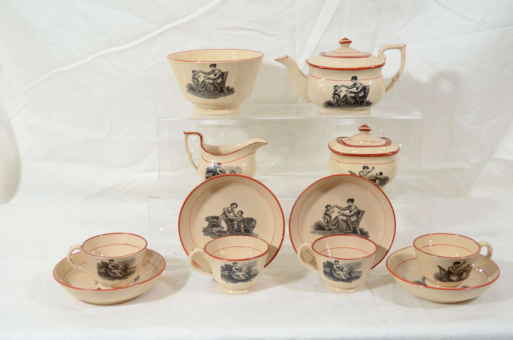 A child's Drabware tea set with Adam Buck transfer prints of mother and daughter each piece outlined in red. The set contains a teapot, sugar box, creamer, slop bowl, and 4 cups and saucers.