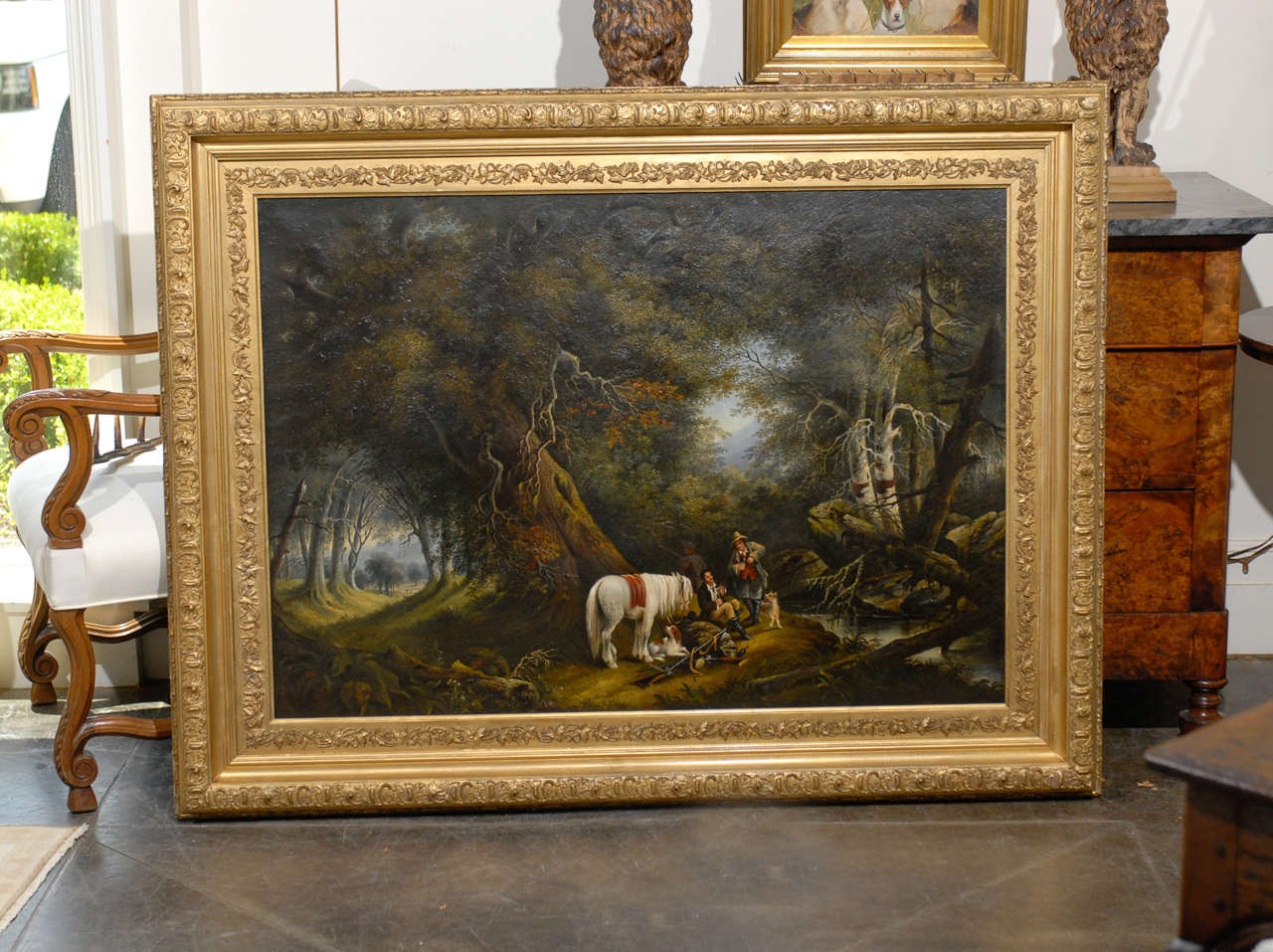 A large horizontal oil on canvas painting with gilded frame featuring men, horses and dogs painted in the style of Alvan Fisher from the Turn of the century. This stunning painting depicts two men resting with their horse, dogs and muskets near a