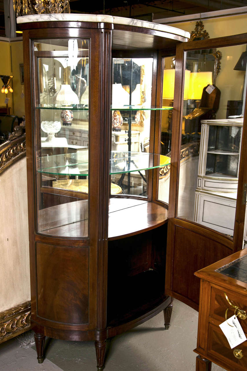 French walnut curio cabinet in Directoire style, circa 1940s, the white beveled half-moon marble atop a glass case with bowed sides opens to glass shelving interior, the bottom with tambour door, raised on fluted tapering legs. By Jansen.