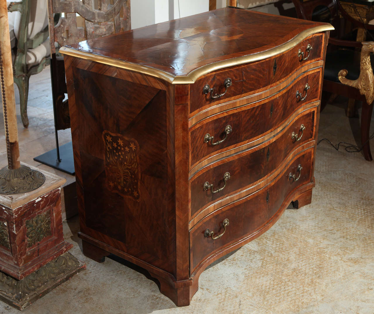 A petite, four-drawer, cross-banded, bow-front, clipped corner commode, with a gilt brass railing. The top features a charming marquetry medallion of a parrot perched on a branch. The sides are embellished with inlaid images of tied bouquets.