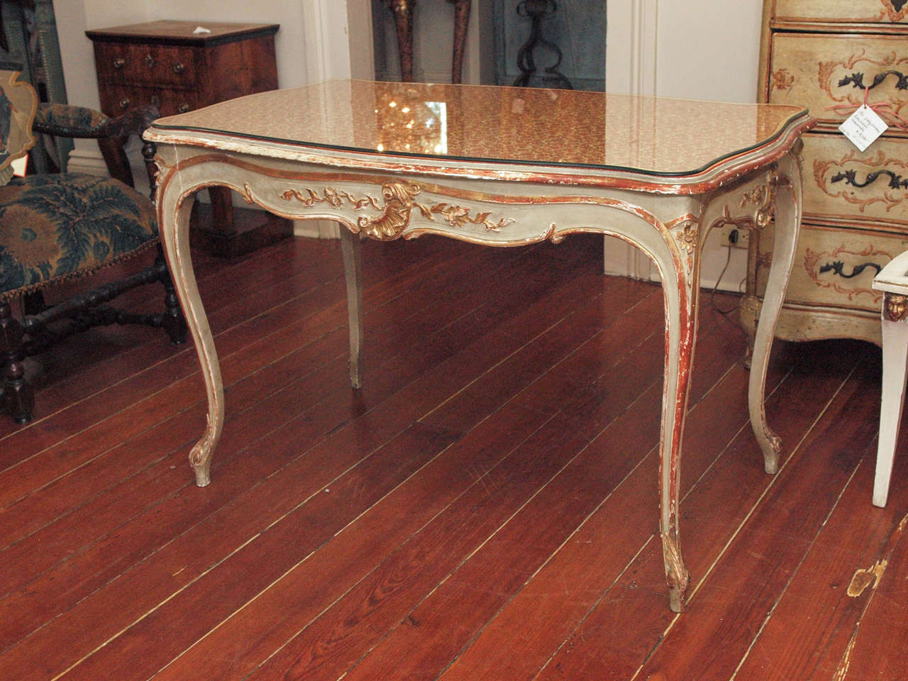 A table or bureau plat in the Louis XV taste, with tall cabriole legs and a drawer, the table with raised, gilded carving and painted,  and having a conforming glass top over fabric.  We imagine the purchaser of this piece replacing the fabric to