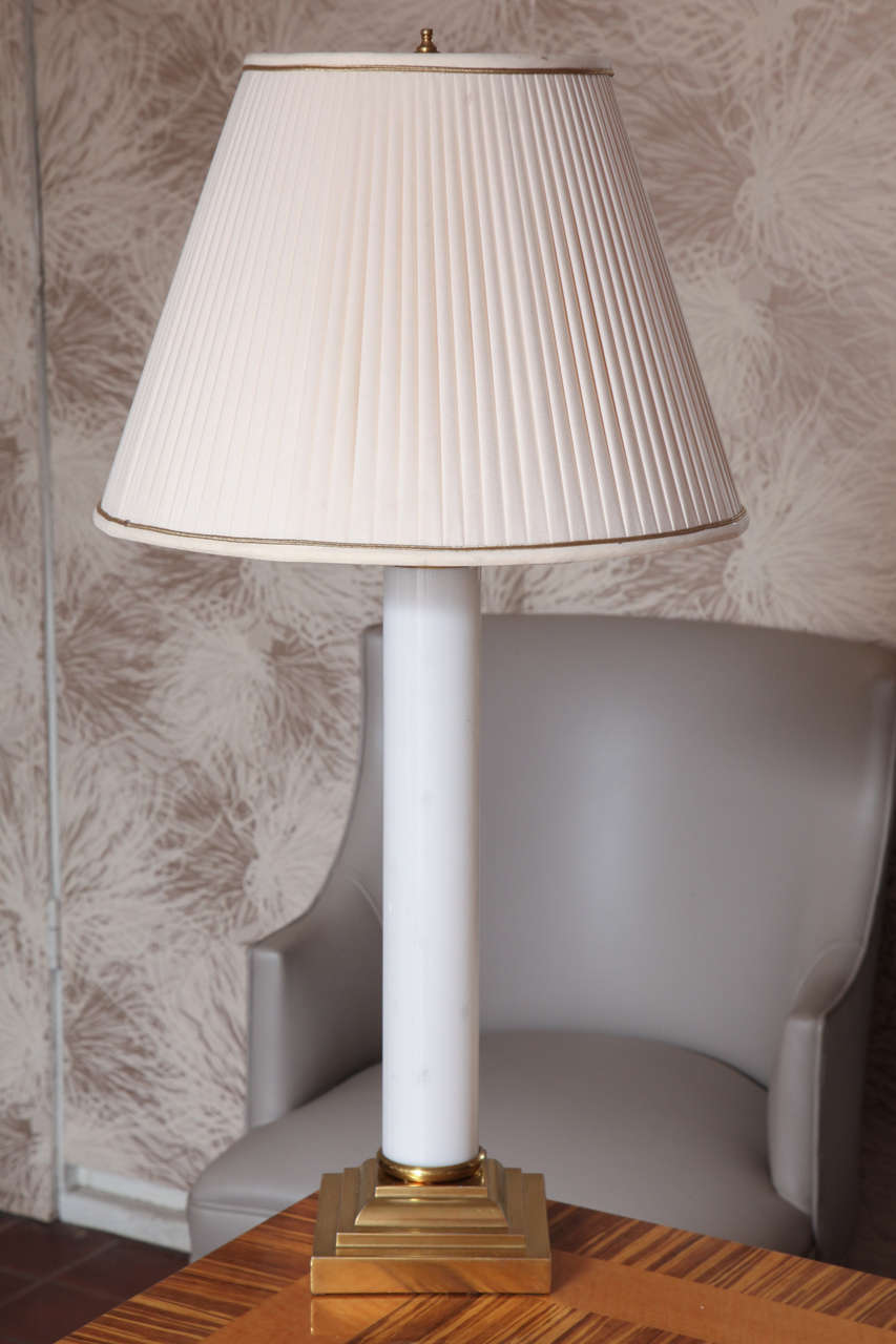 The body of the lamp is a white glossy glass cylinder with a gold brass base. The classic lamp shade is a pleated cream silk fabric with gold trimming. The shade measures 8.5