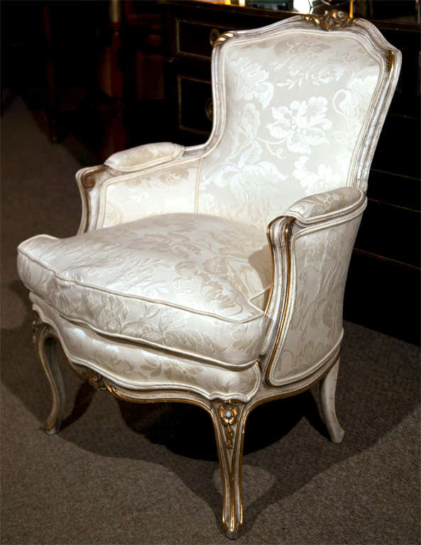 French Louis XV style Bergere chair by Jansen, circa 1940s, distress white painted and parcel-gilt, white floral sateen fabric upholstery, cushioned seat, raised on cabriole legs headed by florette decoration. By Jansen.