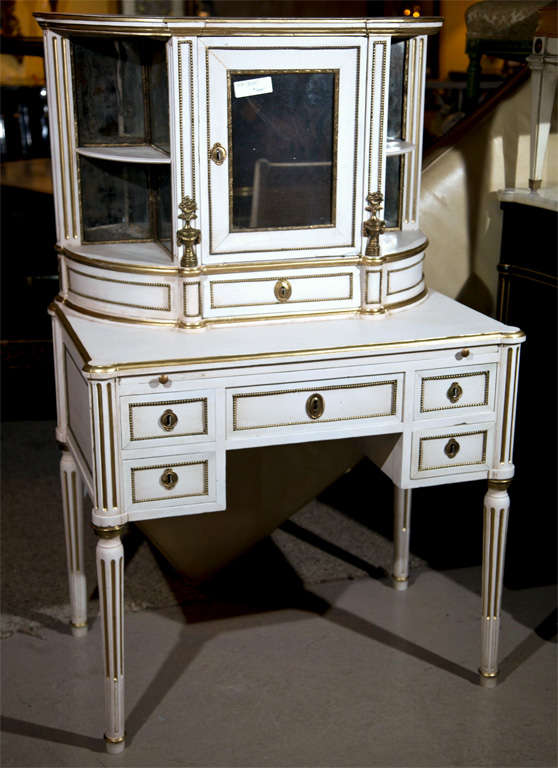 A lovely French Louis XIV style crème peinte and parcel-gilt vanity, circa 1940s, the bow-front top with mirrored door and bronze-mounted finials, over a desk fitted with five banded drawers with brown leather top pull-out surface, raised on fluted