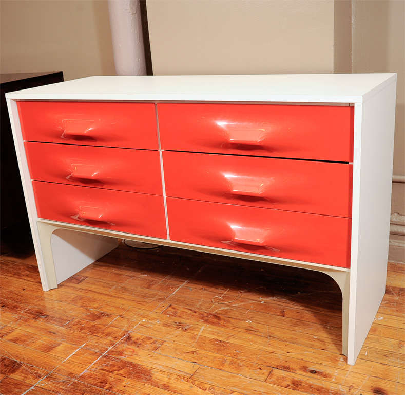 The chest of drawers appears to be from the DF-2000 Series that was created by Raymond Loewy in 1965 and manufactured in France by Doubinsky Freres. It is wood and laminate with solid sides and orange molded plastic drawer fronts. It has three