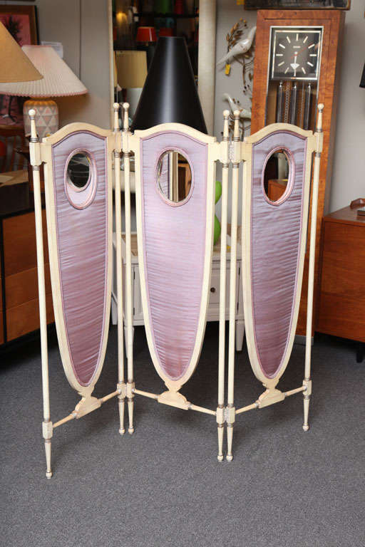 Early 20th century and Edwardian in age, this three panel screen is English Regency Revival in style with its shield shaped panels, upholstered on both sides in silk with bevelled inset oval mirrors on one side. Beautifully painted and silvered