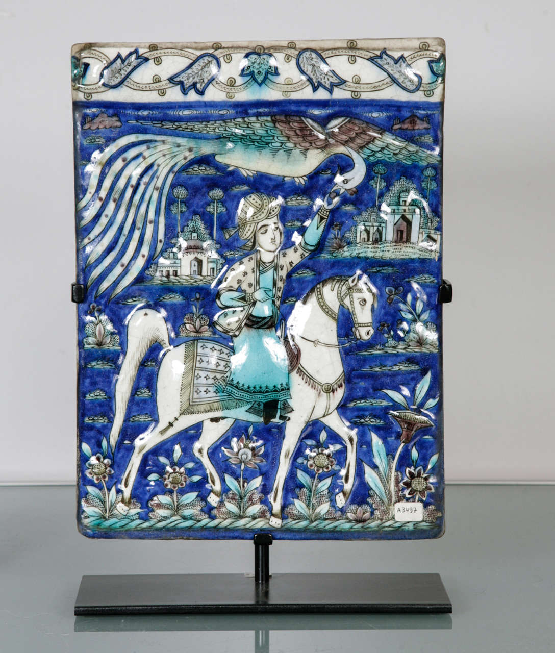 A rectangular polychrome moulded tile depicting an equestrian figure, most likely of a prince - seen extending his arm towards a phoenix-like bird called Huma, symbolic of strength and health. The background cobalt blue with decorative border and