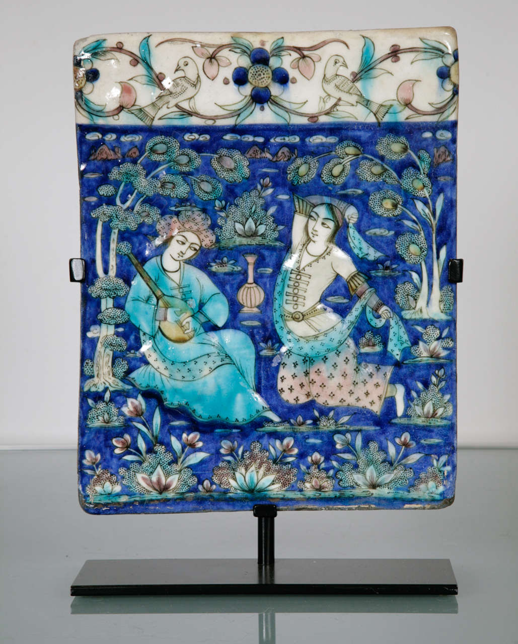 A rectangular polychrome tile depicting a romantic scene with a prince drinking and playing music with a companion. The companion dances with her scarf. Depicted with decorative border and floral landscape.