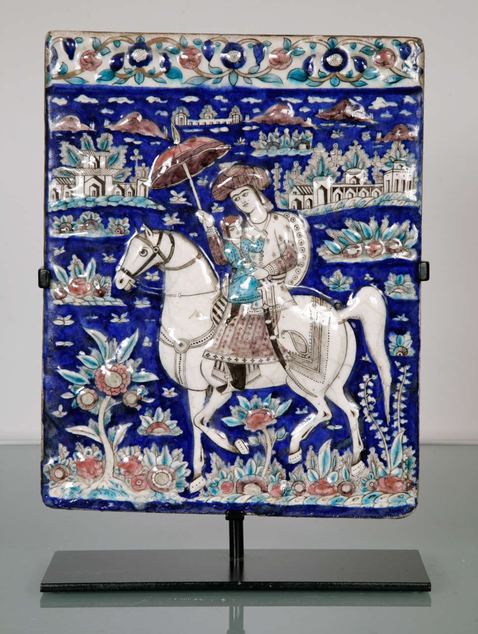 A rectangular polychrome tile depicting an equestrian figure holding a parasol and carrying a child. The background on cobalt blue with decorative border and landscape throughout.
