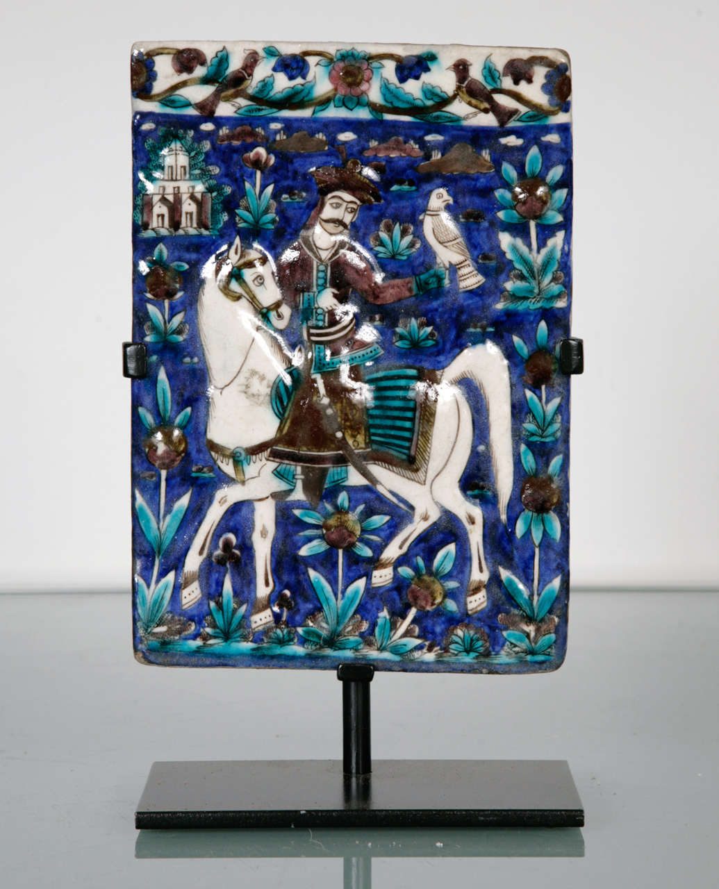 A rectangular polychrome moulded tile depicting an equestrian falconer on a cobalt blue ground with birds and floral border and landscape.