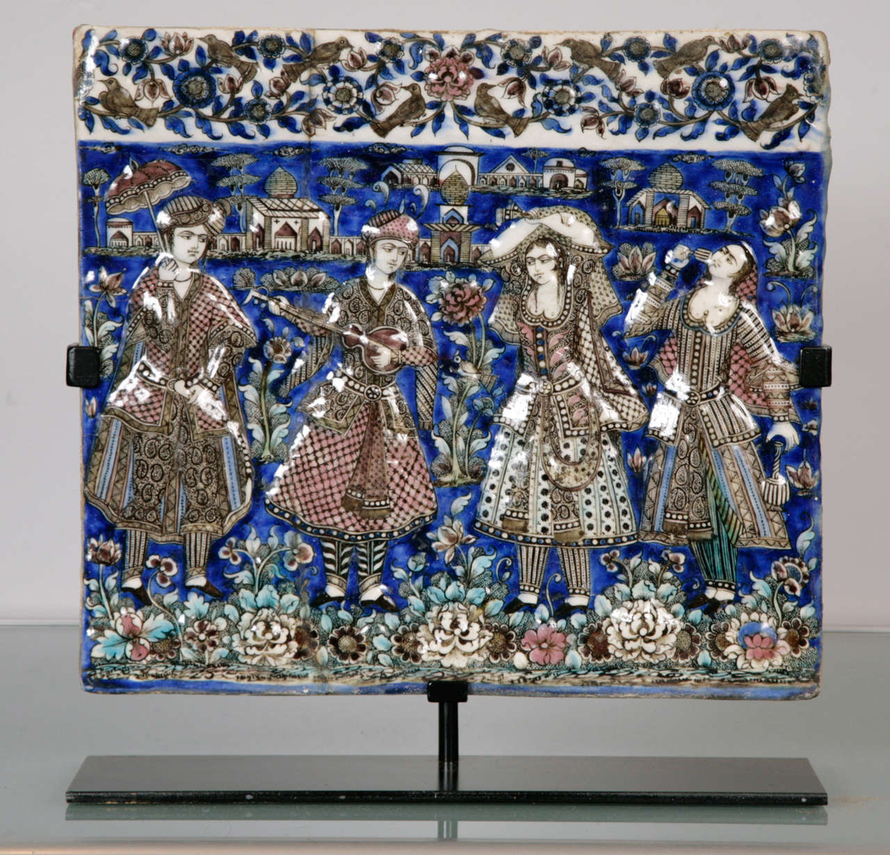 A polychrome tile moulded in relief, the scene depicting musicians and dancers. The figures depicted are dressed in Qajar court costume; the far left figure seen holding a parasol. The musicians are standing in front of a landscape with various