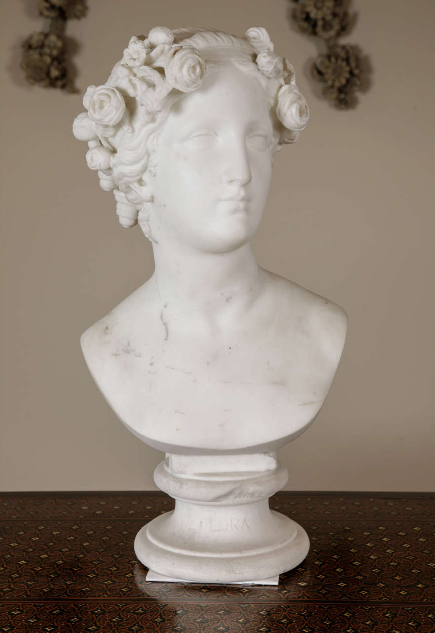 A carved marble bust of Flora, with an elegant and serene countenance. Her hair is gathered to the rear in ringlets, and she is crowned with a garland of roses. Presented on a waisted marble socle. 

In Roman mythology, Flora was the goddess of