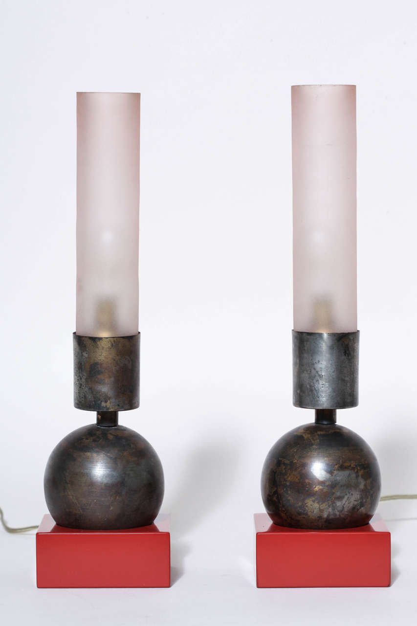Pair of small lamps with a black patinated solid lead spherical foot fitting into a red lacquered wood base and with rose tinted cylindrical ground glass shades. Lacquered wood base 3' on each side. 
Original electrical components with new wiring