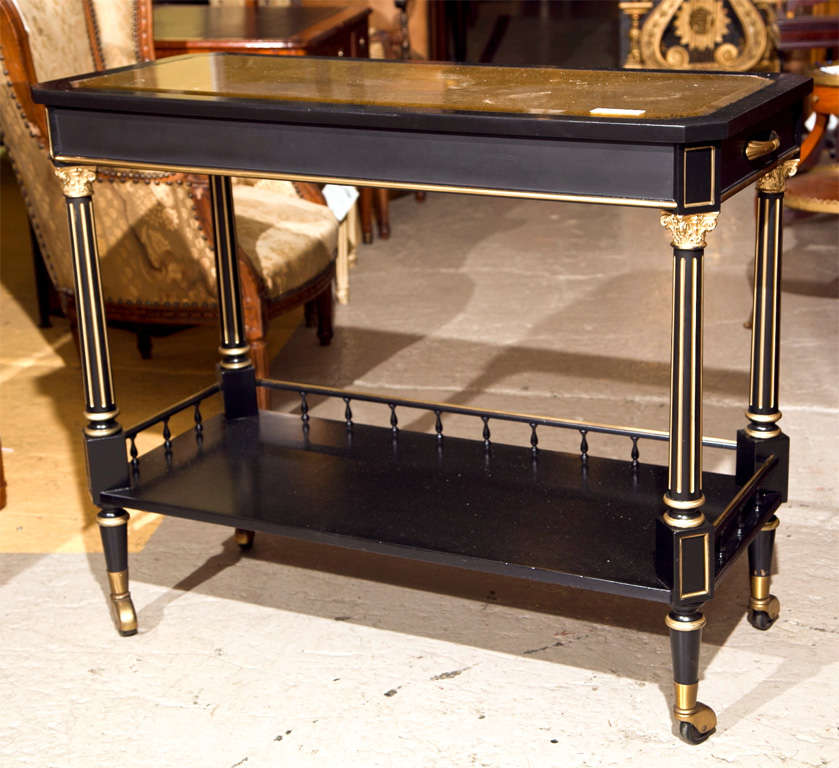 French ebonized and parcel-gilt serving cart, circa 1940s, the top with gilt-glass insert over a narrow frieze with brass handles on each side, supported on four fluted columns, joined by a lower shelf with 3/4 gallery, raised on short bulbous legs