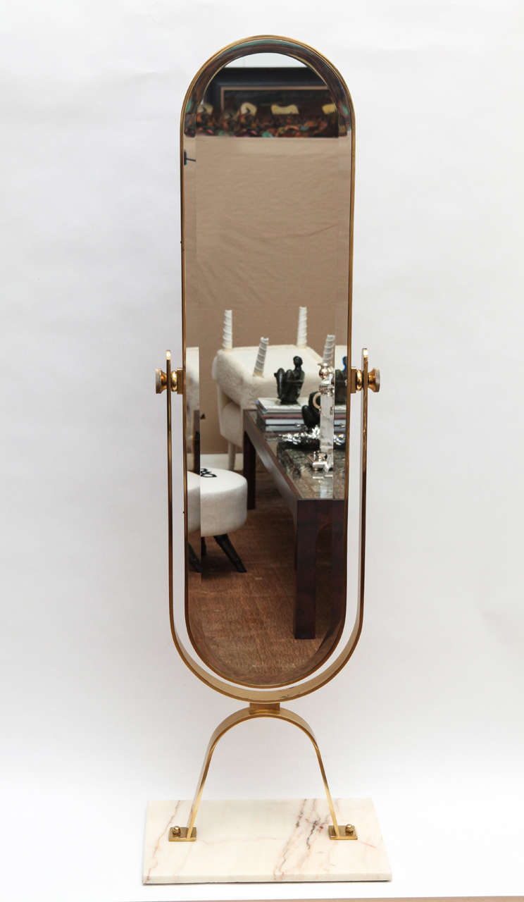 A chic brass framed beveled oval mirror on a pivot. The mirror is set on a pair of brass legs bolted to a 1