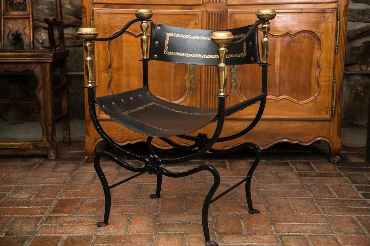 Iron, brass and leather curule armchair. Originally meant for Roman magistrates, this version features tooled and gilded leather on an iron frame with brass mounts joining the back rail to the armrests. The entirety resting on trefoil feet.