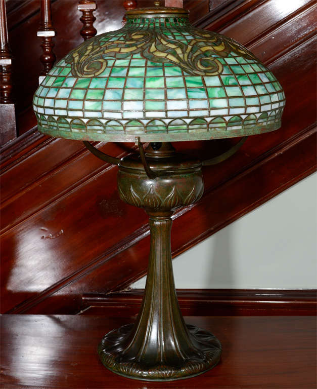 Tiffany Favrile Glass and Bronze Tyler Lamp, 1899-1920
The dome shade in mottled green and white opalescent glass tiles, the upper section with scrolls in mottled golden-amber glass, set on three scrolled arms above a font, the font and base each