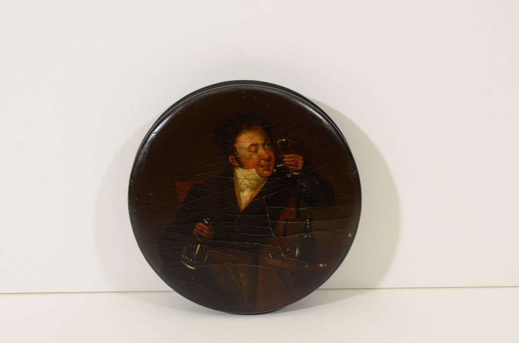 An early 19th century English circular papier-mâché snuff box painted with a gentleman holding a decanter and sizing up a drink