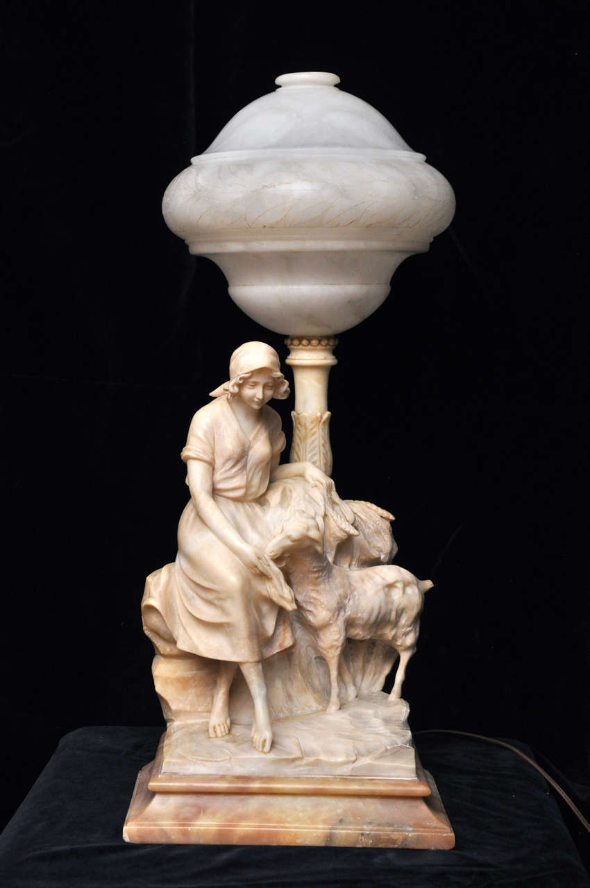 A charming hand carved alabaster figurine lamp, showing a young maiden dressed girl, seated outdoors feeding a lamb.  The young barefooted girl has a scarf covering her hair and she is wearing a European farm dress; her left arm is leaning on shafts