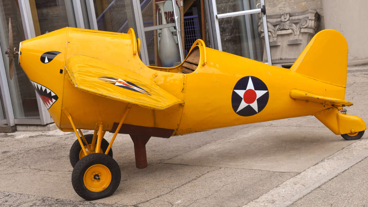 A whimsical mid-20th century canary yellow American carnival fighter plane with wonderfully worn wooden seat, wooden propeller and shark-face nose art.