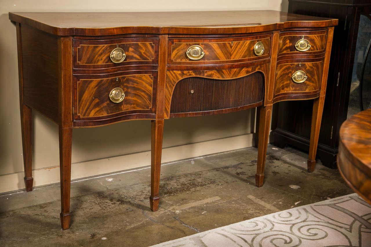 English mahogany double serpentine sideboard, c. 1790-1800, the case with one flanking cupboard and one deep drawer and the center with one thin drawer over a tambour front cabinet. The sideboard exhibits satinwood and ebony string inlay throughout