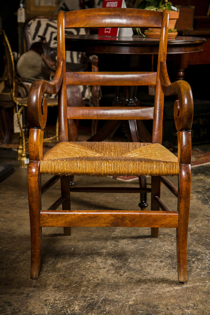 French fruitwood armchair, c. 1840, in the country ladderback style with a rush seat, boldly rolled arms, and saber legs.