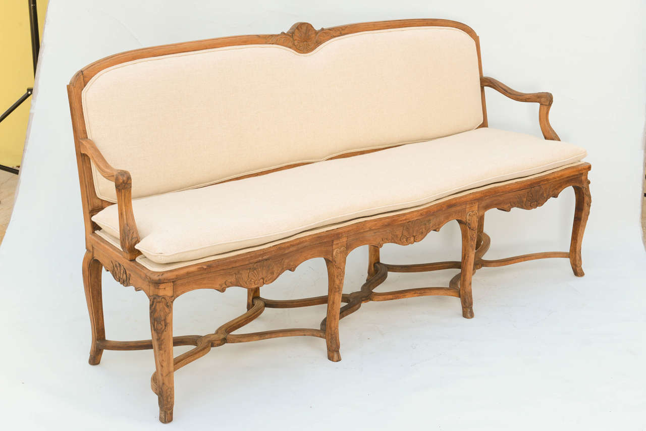 Settee, in Regence style, having a walnut frame; its crestrail carved with stylized scallop shell flanked by scrolls, curving armrests raised on downswept terminals, knife-edge cusion, raised on similarly carved serpentine apron, raised on cabriole