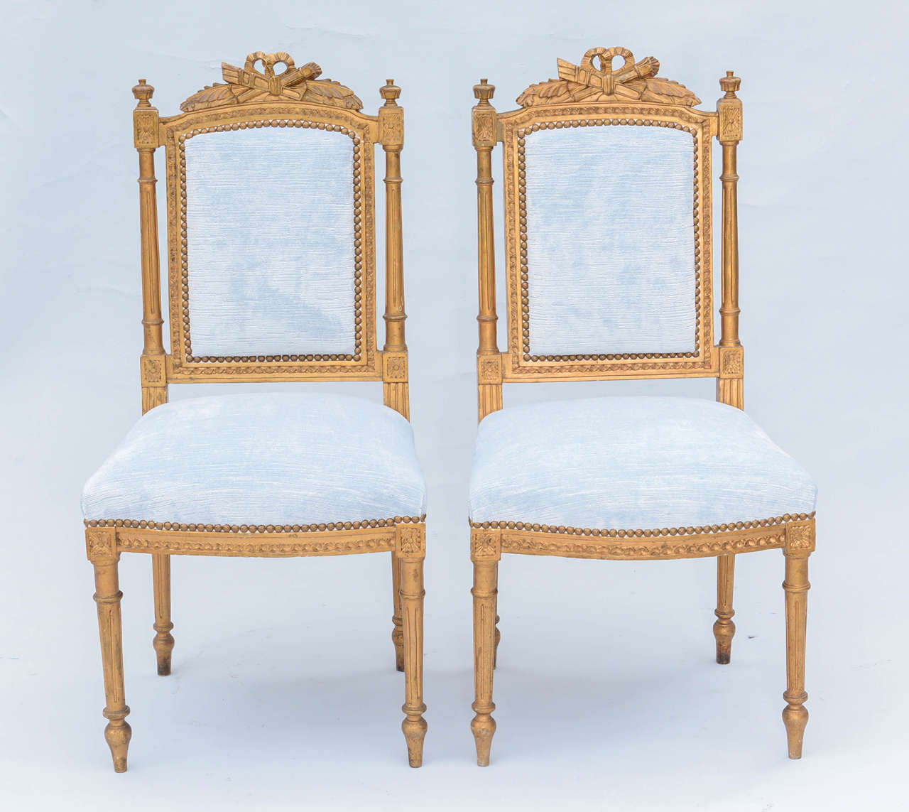 Pair of petite ballroom side chairs, in Louis XVI style; each having a giltwood frame, intricatedly carved crestrail, padded back flanked by fluted stiles, stuff over seat, on bowed apron, raised on round stop-fluted legs, ending in touipe feet.