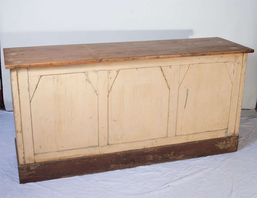 A pine shop counter featuring a panelled base in original distressed off-white and blue paint beneath a polished pine top. A single shelf to the interior.