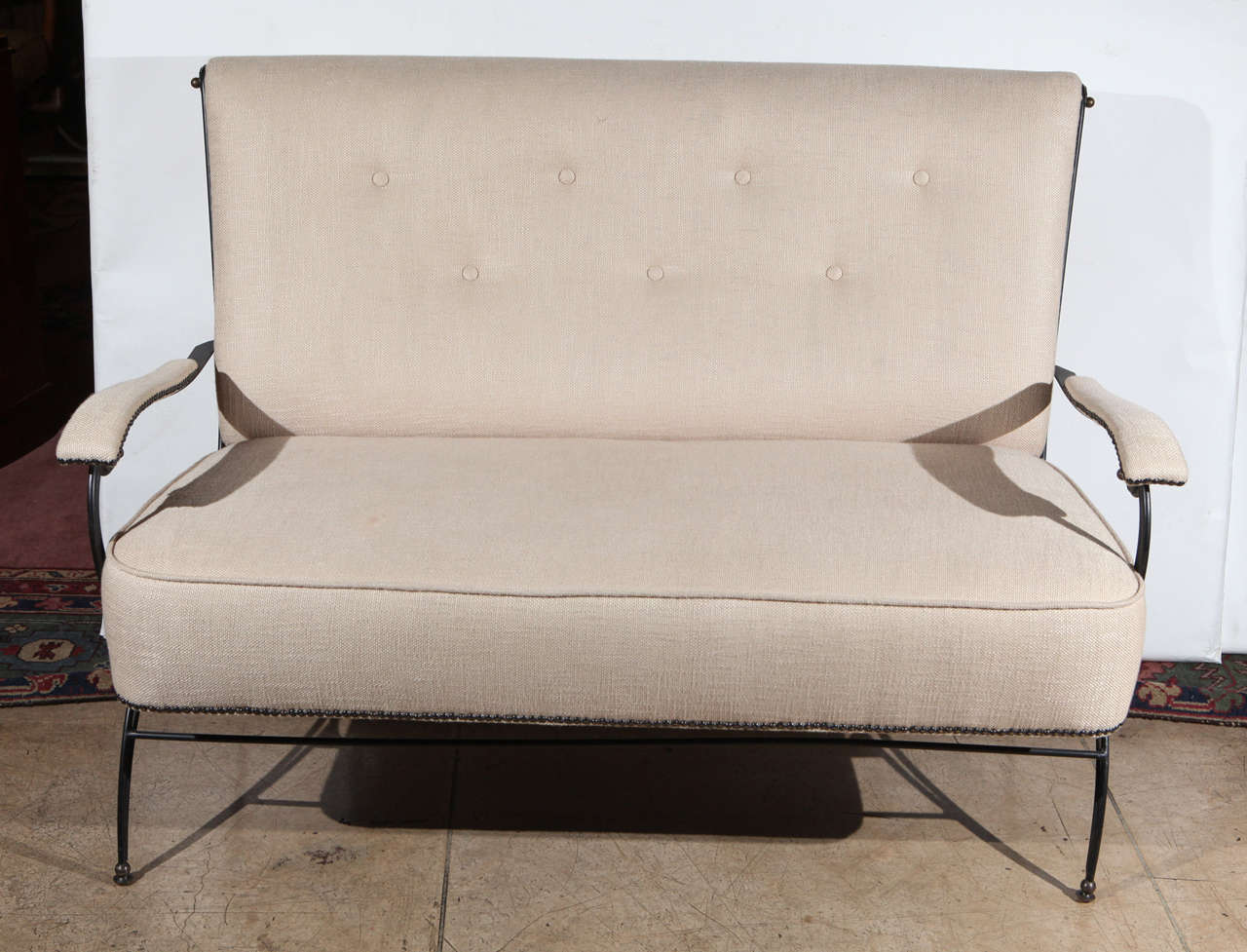 A graceful 1950s French settee in black wrought iron with brass details attributed to Jacques Adnet. Settee is newly upholstered in a cream colored chenille with black nail head details on arms and seat.