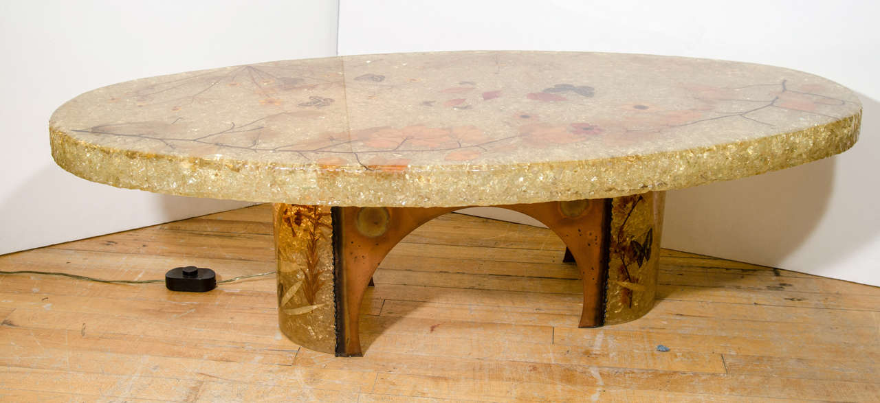 A vintage oval coffee or cocktail table with an amber resin surface and interior lighting. The surface has embedded nature specimens such as leaves, flowers and butterflies.