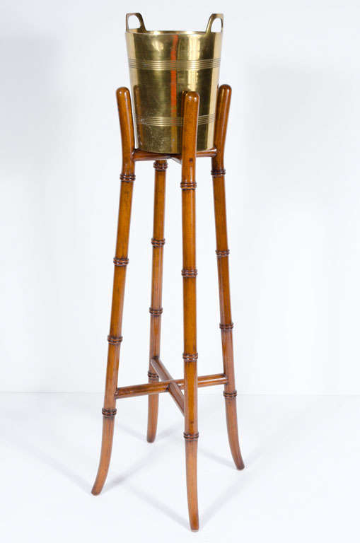 A brass ice bucket with concentric circle band decoration and integral handles supported by a tall faux bamboo wooden stand.  With label 