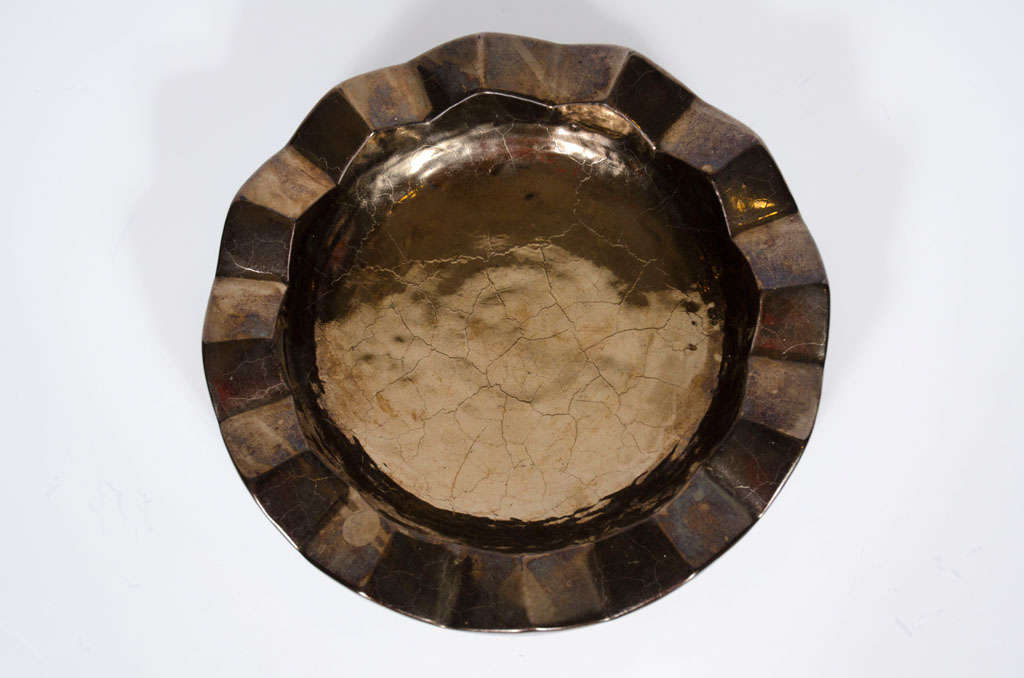 A shimmering shallow ceramic dish in a circular form with a thick and wide zig-zag lip and a gunmetal crackle lustre ware glaze.  Impressed mark to underside.  By Design Technics. American, circa 1950.
