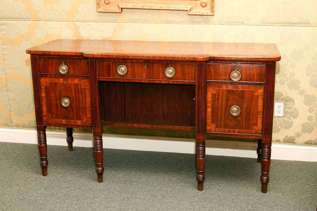 Crossbanded top over two short and one long frieze drawers, above a tambour door compartment flanked by deep drawers separated by reeded columns, raised on ring-turned legs.