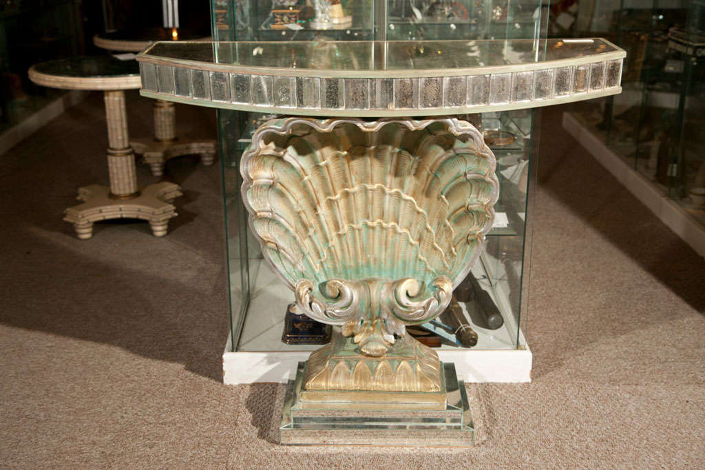Hollywood Regency style console table, circa 1940s, overall painted and parcel-gilt, the half-moon top with antiqued glass veneer as well as glass panels on the frieze. Supported by a shell form pedestal, raised on several layered rectangular