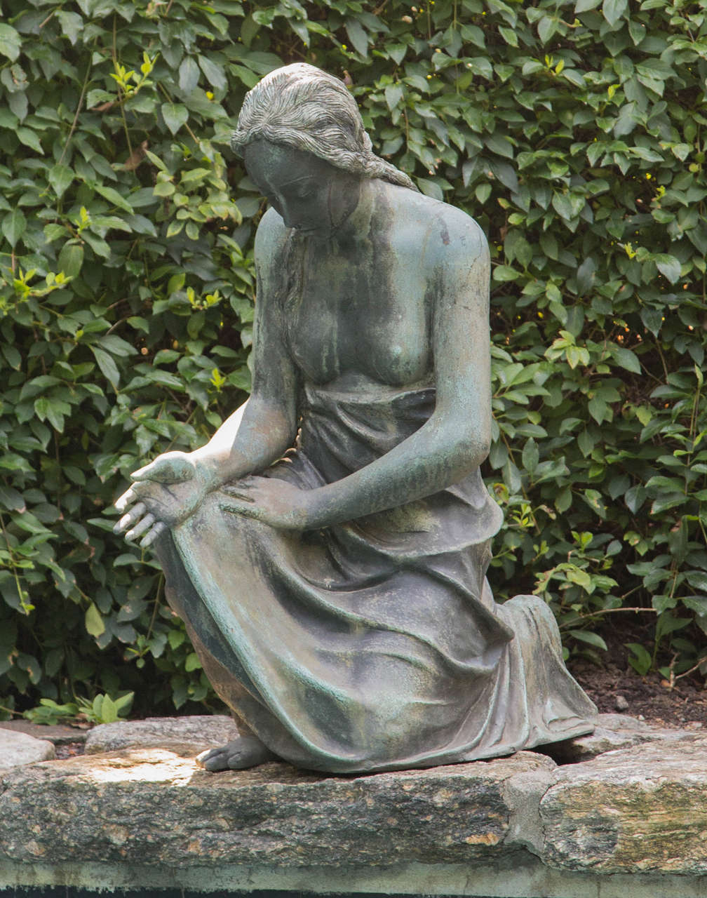An unusual bronze figure in the Arts and Crafts style depicting a partially draped young woman kneeling on one knee with right hand open. The figure, with downward, reflective gaze, is meant to be placed at the edge of a pool.