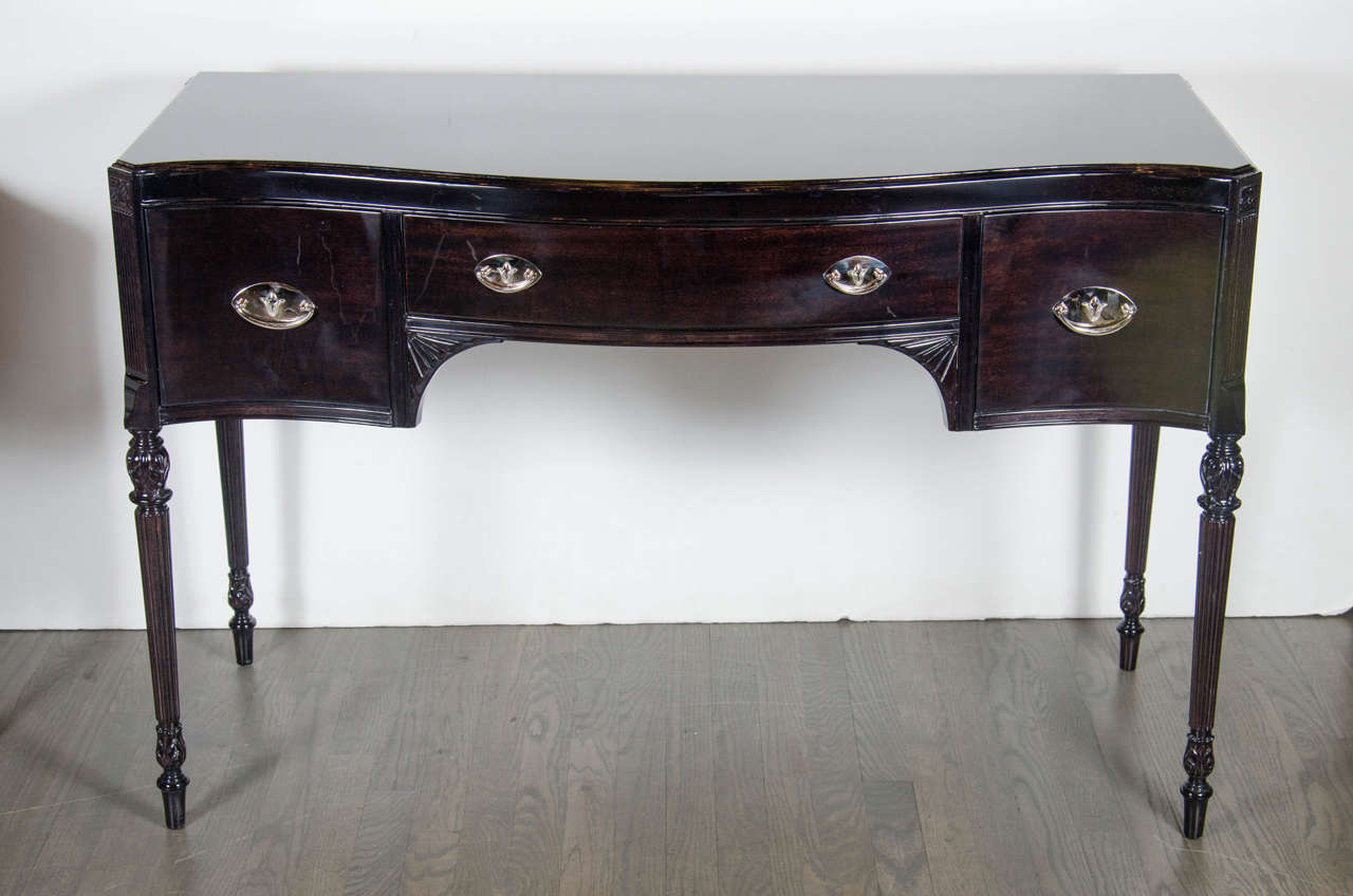 This stunning vanity or desk features a bowed front design with stylized balustrade form legs and stylized hand carved fan detailing. It also in made of ebonized mahogany with polished nickeled pulls. It has three generous sized drawers for great