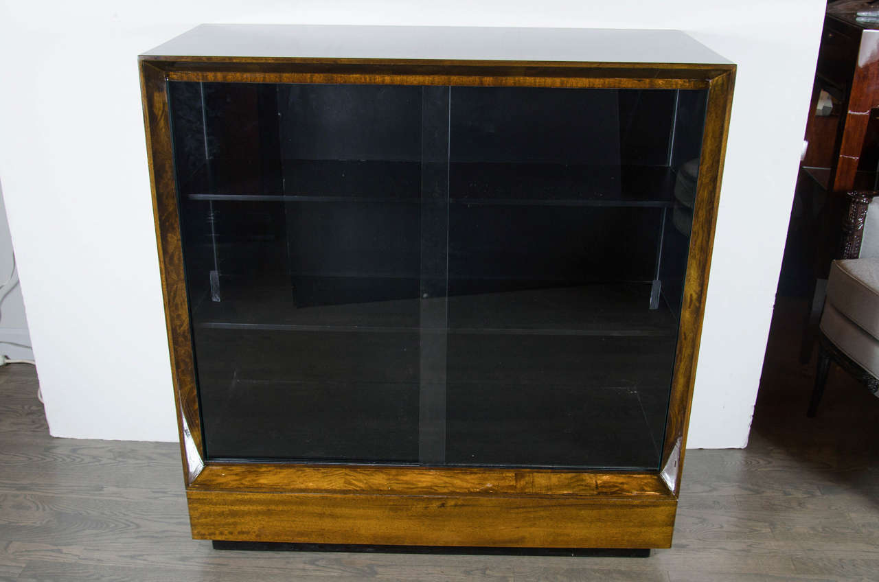 This stunning Gilbert Rohde's Art Deco bookcase in book-matched paldao wood has an ebonized interior and black lacquered base. The bookcase has two sliding glass doors and two large shelves. This piece has been restored to mint condition. It is also