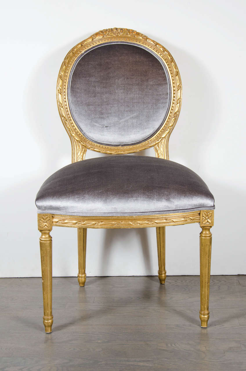 Set of 12 stunning Louis XIV style dining chairs that have been gilded in 24-karat gold-leaf. They feature rounded backs with hand carved detailing of a beaded border topped with a foliage design and upholstered in a chic silk velvet in colorings of