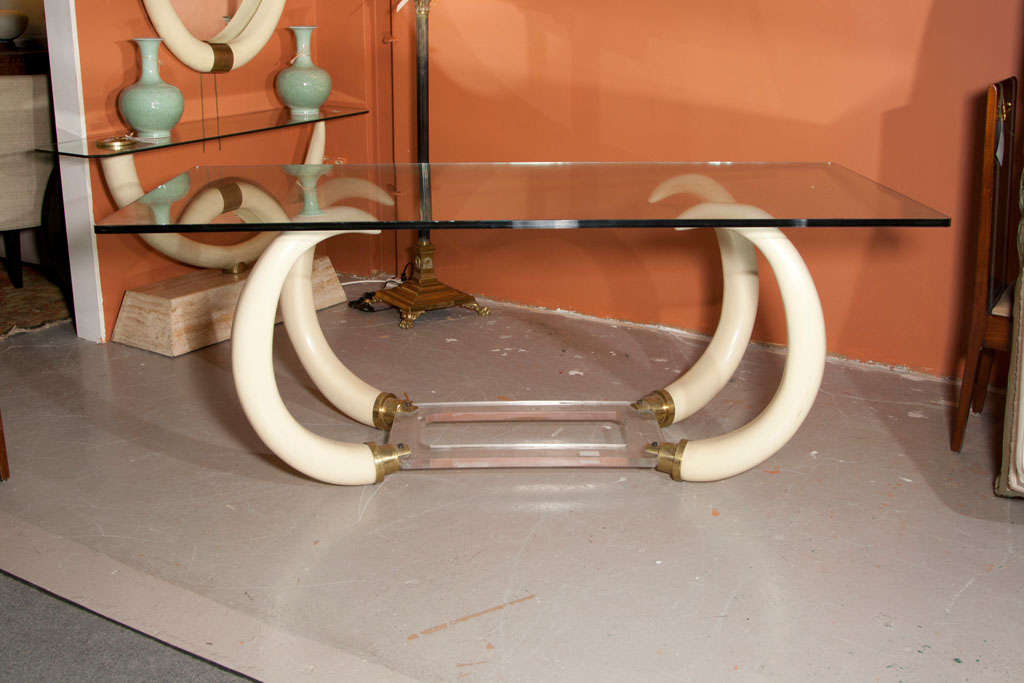 An intriguing dining table, circa 1960s, the thick glass top over a base decorated with four faux elephant tusks joint by a rectangular lucite stretcher.